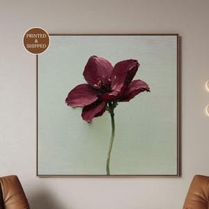 May include: A square framed wall art piece featuring a single dark burgundy flower with thick, textured impasto paint strokes centred on a pale sage green background. The artwork is displayed in a thin wooden frame with a circular brown logo.