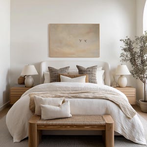 May include: A bedroom scene featuring a bed with white bedding, decorative pillows, and a woven bench at the foot. Two bedside tables with lamps flank the bed. A framed artwork hangs above the bed, and a potted plant is in the corner.