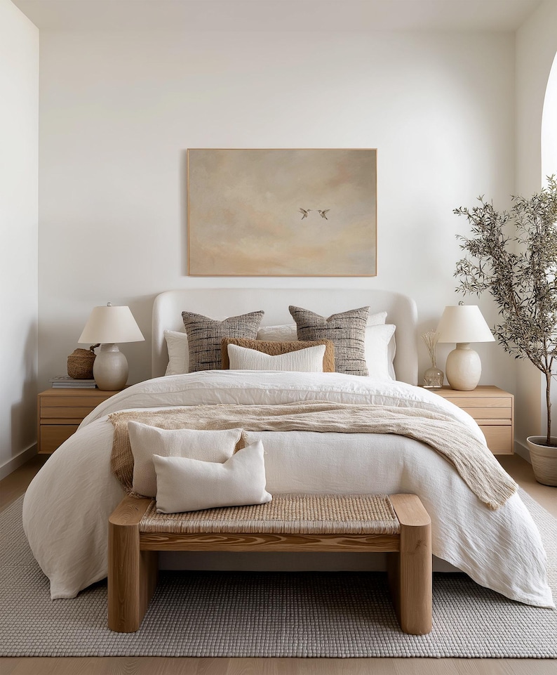 May include: A bedroom scene featuring a bed with white bedding, decorative pillows, and a woven bench at the foot. Two bedside tables with lamps flank the bed. A framed artwork hangs above the bed, and a potted plant is in the corner.