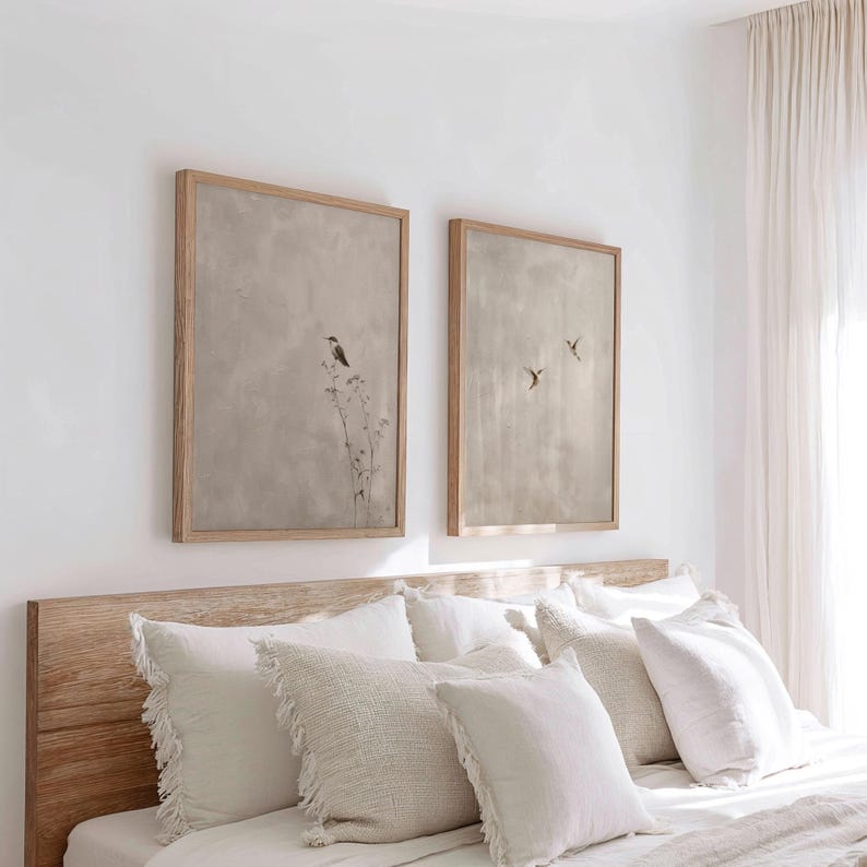 Op de afbeelding: Twee ingelijste kunstwerken met een neutrale kleurenpalet. E&eacute;n toont een vogel op een plant, de andere toont drie vogels in vlucht. De kunstwerken worden weergegeven boven een bed met witte kussens en een houten hoofdbord.