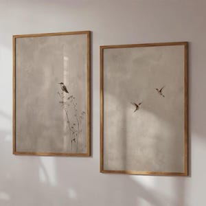 Op de afbeelding: Twee ingelijste kunstdrukken met een neutraal kleurenpalet. E&eacute;n print toont een vogel op een plant, de andere toont drie vogels in vlucht. De lijsten hebben een warme houttint, die de zachte, gedempte achtergrond aanvult.