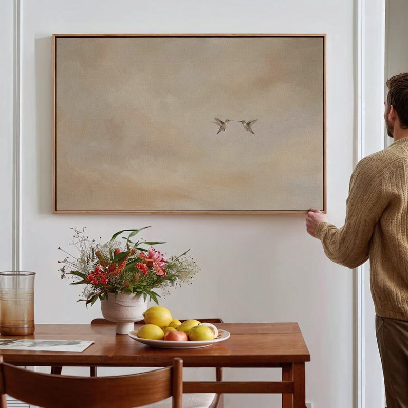 May include: A framed rectangular artwork with a light brown wooden frame. The artwork depicts two birds in flight against a neutral background. The painting is being hung on a white wall. A wooden table is in the foreground, with a vase of flowers and a plate of fruit.