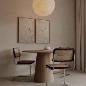 Op de afbeelding: Een eethoek met een ronde houten tafel en drie stoelen met geweven rugleuningen en zittingen. Twee ingelijste kunstwerken hangen aan de muur. Boven de tafel hangt een grote, ronde lamp. De kamer heeft een neutraal kleurenpalet.