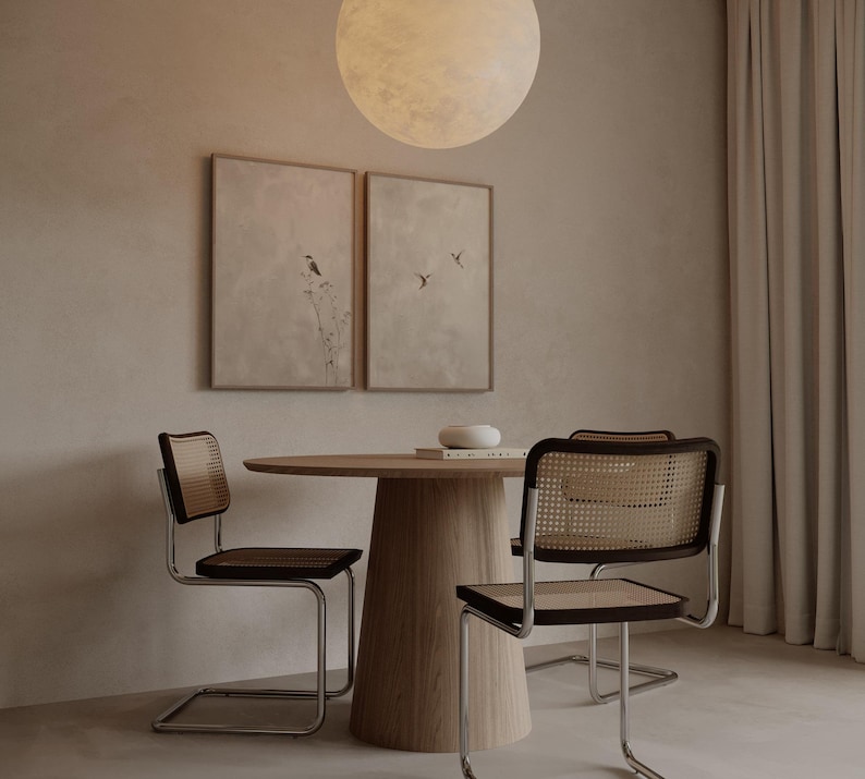 Op de afbeelding: Een eethoek met een ronde houten tafel en drie stoelen met geweven rugleuningen en zittingen. Twee ingelijste kunstwerken hangen aan de muur. Boven de tafel hangt een grote, ronde lamp. De kamer heeft een neutraal kleurenpalet.