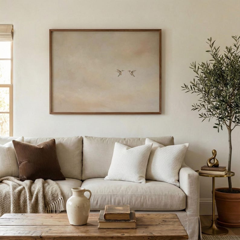 May include: A living room scene featuring a beige sofa with various throw pillows in white and brown. A wooden coffee table holds books and a vase. A framed artwork with birds hangs above the sofa, and a small olive tree is to the right.