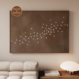 May include: A framed wall art print featuring a flock of white birds in flight against a brown background. The artwork has a simple, minimalist design and is displayed in a room with a neutral color palette. The text "PRINTED & SHIPPED" is in a circle in the upper left corner.