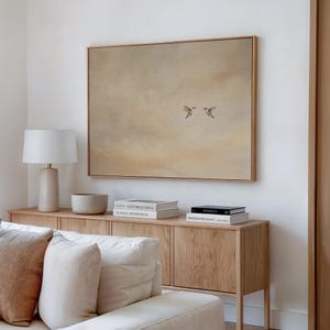 May include: A framed artwork featuring two birds in flight against a muted beige background. The artwork is displayed above a light wood cabinet with a lamp, a bowl, and stacked books. The scene evokes a sense of calm and natural beauty.