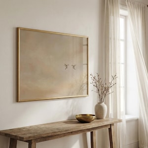May include: A framed artwork with a gold border features a soft, muted landscape with two birds in flight. The artwork hangs above a rustic wooden console table. A white vase with branches and a gold bowl are on the table.