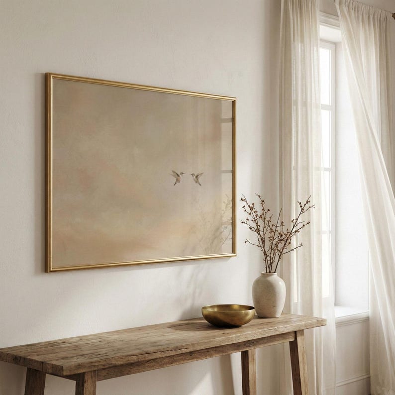 May include: A framed artwork with a gold border features a soft, muted landscape with two birds in flight. The artwork hangs above a rustic wooden console table. A white vase with branches and a gold bowl are on the table.