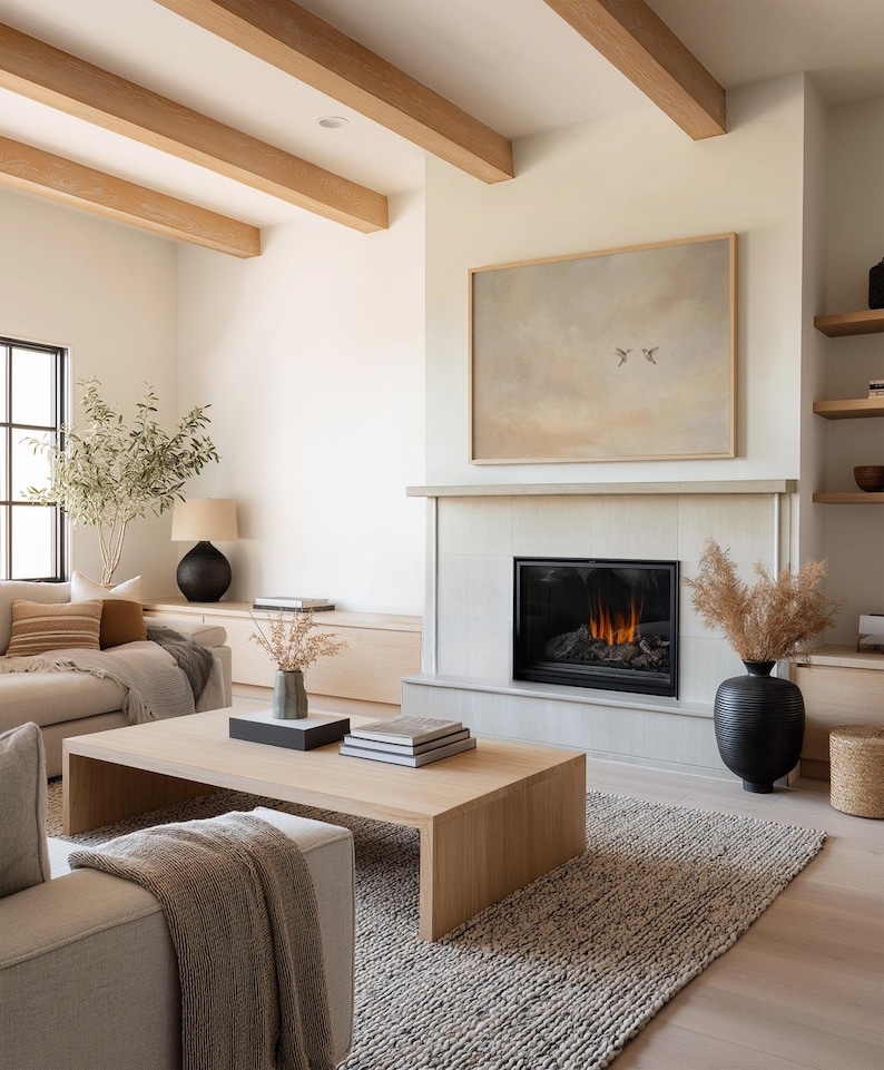 May include: A modern living room with a neutral color palette. Features include a light wood coffee table, a fireplace with a painting above, and a textured rug. The room is decorated with a black vase, books, and a sofa with throw pillows.