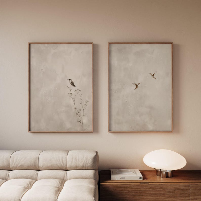 Set of 2 minimalist bird wall art prints in light wooden frames, featuring soft neutral textured backgrounds with delicate birds and subtle botanical elements, styled above a beige couch in a calm Scandinavian living room interior.