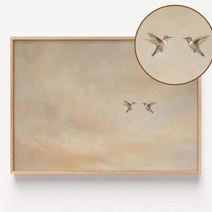 May include: A framed art print featuring two hummingbirds in flight, set against a soft, muted background of beige and tan hues. A circular inset at the top right shows a close-up of two hummingbirds. The artwork is encased in a light brown wooden frame.