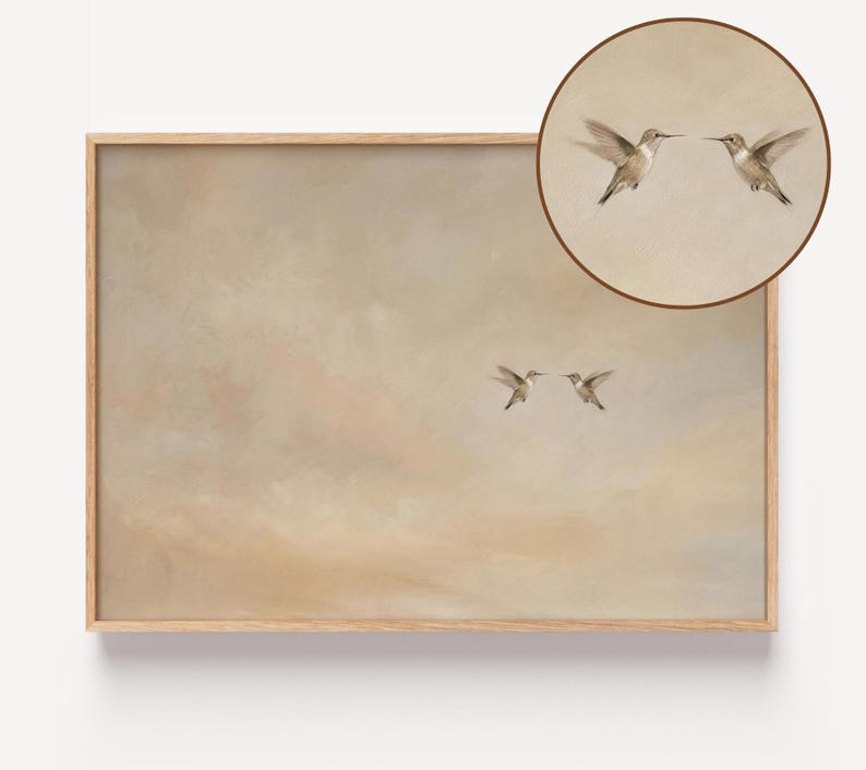 May include: A framed art print featuring two hummingbirds in flight, set against a soft, muted background of beige and tan hues. A circular inset at the top right shows a close-up of two hummingbirds. The artwork is encased in a light brown wooden frame.