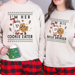 Może przedstawiać: Dwie pasujące beżowe bluzy z napisami "I'M HER COOKIE EATER" i "I'M HIS MILK DRINKER". Bluzy mają kreskówkowe grafiki mleka i ciasteczek. Modele mają na sobie czerwono-czarne spodnie od piżamy w kratę.