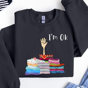 Może przedstawiać: Czarna bluza z białym nadrukiem przedstawiającym rękę wystającą z kupy kolorowych tkanin. Nad ręką widnieje napis "I'm OK".