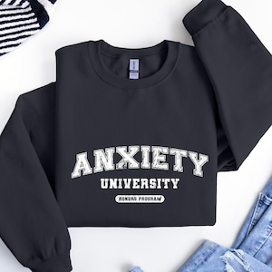 May include: A dark gray sweatshirt with the text "Anxiety University Honors Program" in white. The text is in a vintage collegiate style font.