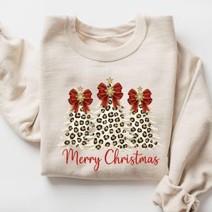 May include: A cream-coloured sweatshirt featuring three Christmas trees with leopard print patterns. Each tree has a red bow and a gold star. The words "Merry Christmas" are written in red script below the trees.