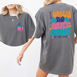 Op de afbeelding: Donkergrijs t-shirt met een kleurrijke afbeelding op de achterkant met de tekst "I ONLY DO BUTT STUFF AT THE GYM". De voorkant heeft een kleine perzikafbeelding. Het shirt heeft korte mouwen en een relaxte pasvorm.