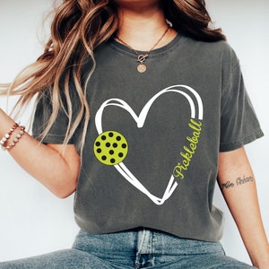 May include: A gray t-shirt with a white heart design featuring a green and black pickleball. The text "Pickleball" is written inside the heart.