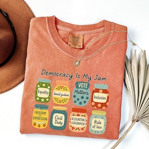 Pode incluir: Camiseta coral de manga comprida com a frase "Democracy Is My Jam" e ilustrações de potes com palavras como "Equality" e "Vote Matters". Um colar de ouro e um chapéu castanho também são visíveis.