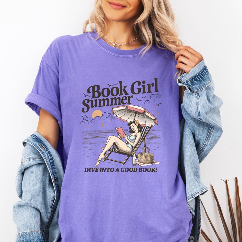 May include: A purple t-shirt with the words "Book Girl Summer" and a graphic of a woman reading on a beach. The graphic includes a beach umbrella, a beach chair, and the text "DIVE INTO A GOOD BOOK!"