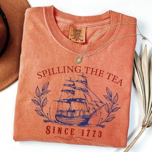 May include: Coral-colored t-shirt with the phrase "Spilling the Tea Since 1773" and a blue illustration of a sailing ship. A gold necklace with a pendant rests on the shirt. The shirt has a crew neck and short sleeves.