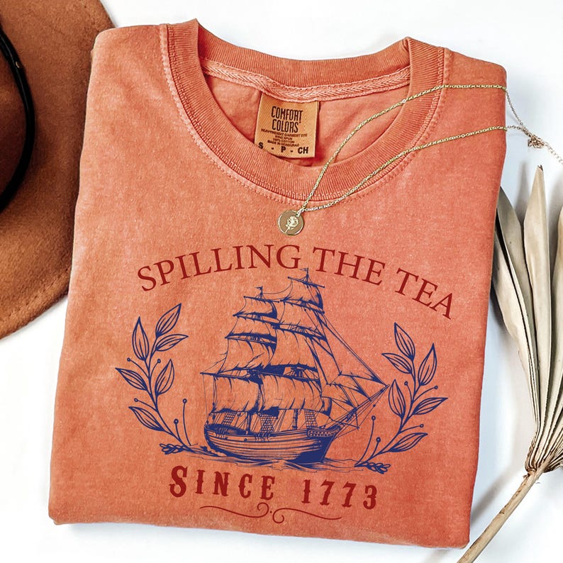 May include: Coral-colored t-shirt with the phrase "Spilling the Tea Since 1773" and a blue illustration of a sailing ship. A gold necklace with a pendant rests on the shirt. The shirt has a crew neck and short sleeves.