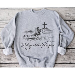 May include: A grey sweatshirt featuring a black and white illustration of a person riding a horse towards a cross. The text "Riding with Purpose" and "Proverbs 19:21" is printed on the sweatshirt.