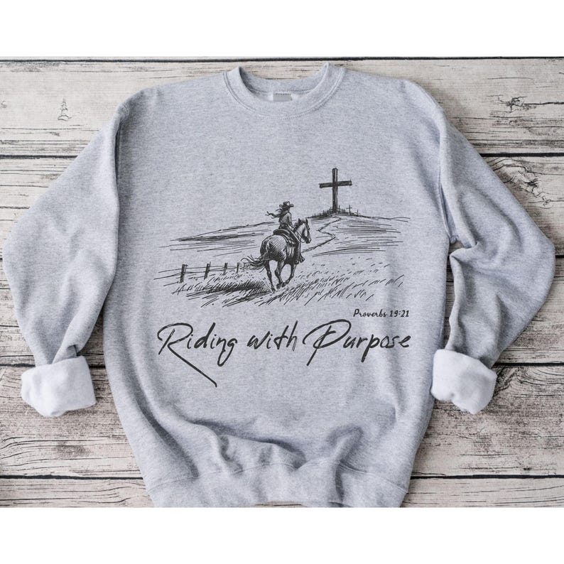 May include: A grey sweatshirt featuring a black and white illustration of a person riding a horse towards a cross. The text "Riding with Purpose" and "Proverbs 19:21" is printed on the sweatshirt.