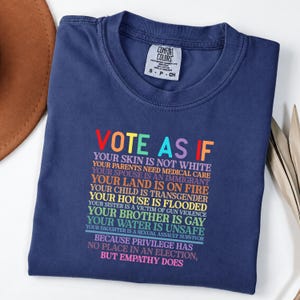 May include: A navy blue crew neck t-shirt featuring multi-colored text that reads: VOTE AS IF YOUR SKIN IS NOT WHITE, YOUR PARENTS NEED MEDICAL CARE, YOUR SPOUSE IS AN IMMIGRANT, YOUR LAND IS ON FIRE, YOUR CHILD IS TRANSGENDER, YOUR HOUSE IS FLOODED.