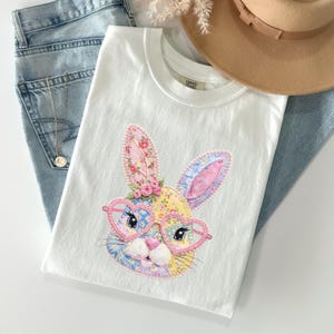 May include: A white t-shirt featuring a colorful patchwork-style rabbit graphic. The rabbit has long ears with floral patterns, a yellow face, and wears pink heart-shaped glasses. The design includes small pink roses near the rabbit's ear.