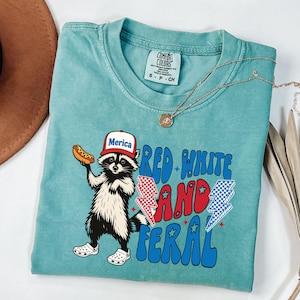 May include: A turquoise Comfort Colors t-shirt featuring a raccoon wearing a "Merica" hat and holding a hot dog. The shirt has the text "Red White and Feral" with lightning bolt graphics. A gold necklace is draped across the shirt.