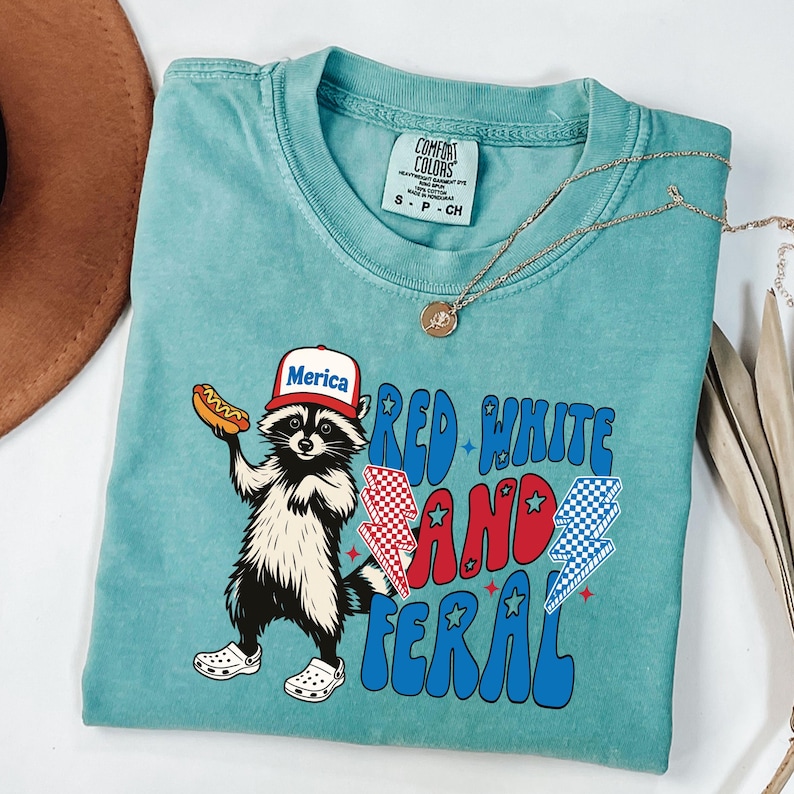 May include: A turquoise Comfort Colors t-shirt featuring a raccoon wearing a "Merica" hat and holding a hot dog. The shirt has the text "Red White and Feral" with lightning bolt graphics. A gold necklace is draped across the shirt.