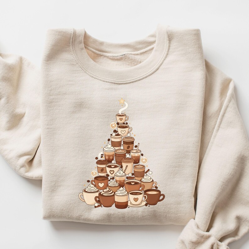 May include: Beige sweatshirt featuring a coffee cup Christmas tree design. The tree is composed of various coffee cups with whipped cream, hearts, and coffee beans. A star tops the tree, and steam rises from the top cup.