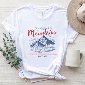 Könnte beinhalten: Weißes T-Shirt mit einem Bergmotiv und dem Text "He who forms the Mountains" in Rot und Blau. Das T-Shirt enthält auch den Bibelvers Amos 4:13.