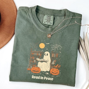 Op de afbeelding: Olijfgroen t-shirt met een afbeelding van een spook dat leest voor een boekenplank, pompoenen en een volle maan. De tekst "Read In Peace" staat eronder. Een zilveren ketting met hanger ligt op het shirt.