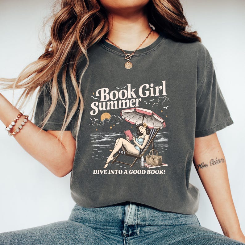 May include: Dark gray t-shirt with a graphic that reads "Book Girl Summer." The graphic depicts a woman reading on a beach chair under an umbrella. The text "Dive into a good book!" is also included.