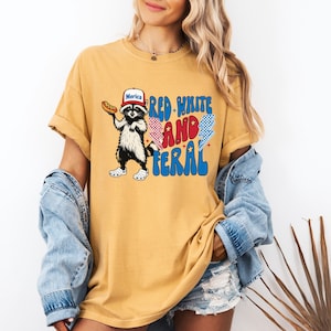 May include: Mustard yellow t-shirt featuring a raccoon wearing a "Merica" hat and holding a hot dog. The shirt has the text "Red, White, and Feral" in a patriotic font. The raccoon is wearing white and black shoes.