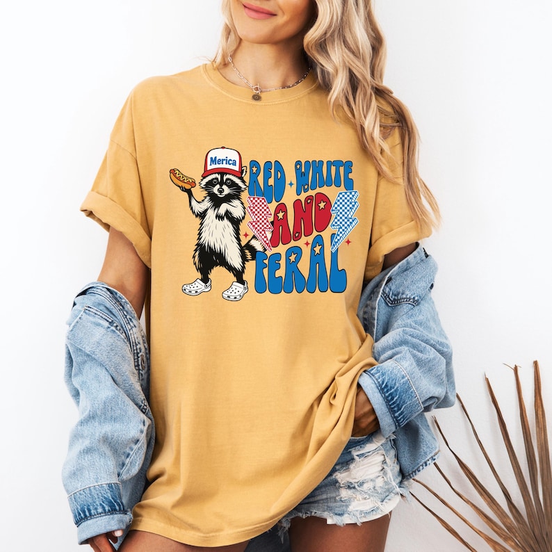 May include: Mustard yellow t-shirt featuring a raccoon wearing a "Merica" hat and holding a hot dog. The shirt has the text "Red, White, and Feral" in a patriotic font. The raccoon is wearing white and black shoes.