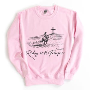 May include: Pink sweatshirt featuring a black and white illustration of a person riding a horse towards a cross. The text "Proverbs 19:21" and "Riding with Purpose" are printed on the sweatshirt.