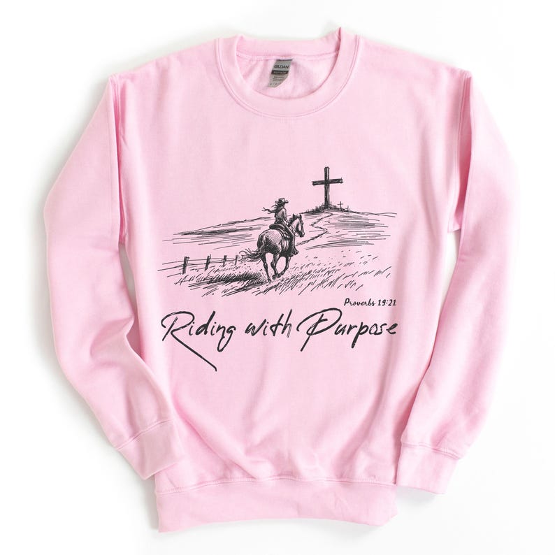 May include: Pink sweatshirt featuring a black and white illustration of a person riding a horse towards a cross. The text "Proverbs 19:21" and "Riding with Purpose" are printed on the sweatshirt.