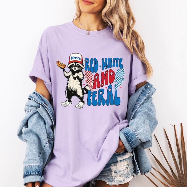 May include: A light purple t-shirt with a graphic of a raccoon wearing a hat that says "Merica" and holding a hot dog. The text "Red, White, and Feral" is printed in red, white, and blue. The raccoon is wearing white shoes.