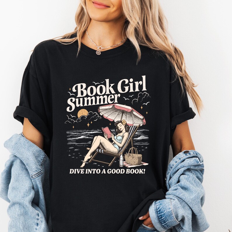 May include: Black t-shirt with a graphic that reads "Book Girl Summer" above an illustration of a woman reading on a beach. The graphic includes a beach umbrella, ocean, and the text "DIVE INTO A GOOD BOOK!"
