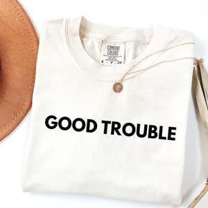 May include: A cream colored t-shirt with the text "GOOD TROUBLE" printed in black. The shirt is folded and laid flat on a white surface.