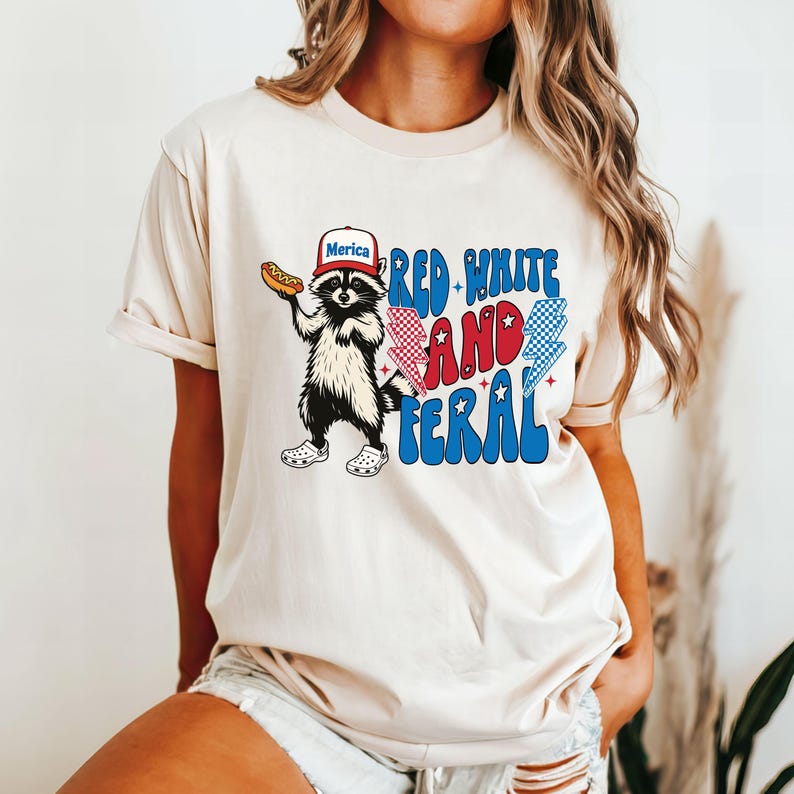 May include: Cream-colored t-shirt featuring a raccoon wearing a "Merica" hat and holding a hot dog. The shirt has the text "Red White and Feral" in red, white, and blue colors, with lightning bolt and star graphics.