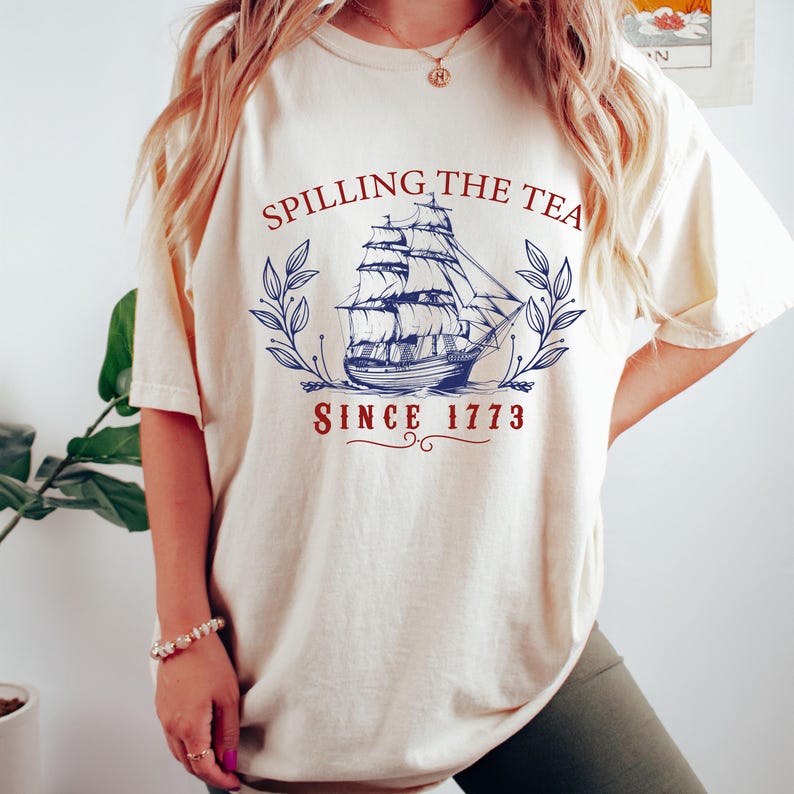 May include: Cream-colored t-shirt featuring a navy blue sailing ship graphic with the text "Spilling the Tea Since 1773" in red and navy blue. The design includes a laurel wreath around the ship, creating a vintage aesthetic.