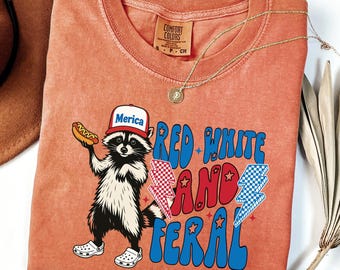 May include: Orange Comfort Colors t-shirt featuring a raccoon wearing a "Merica" hat and holding a hot dog. The shirt has the text "Red White and Feral" in red and blue, with lightning bolt graphics. A gold necklace is draped on the shirt.