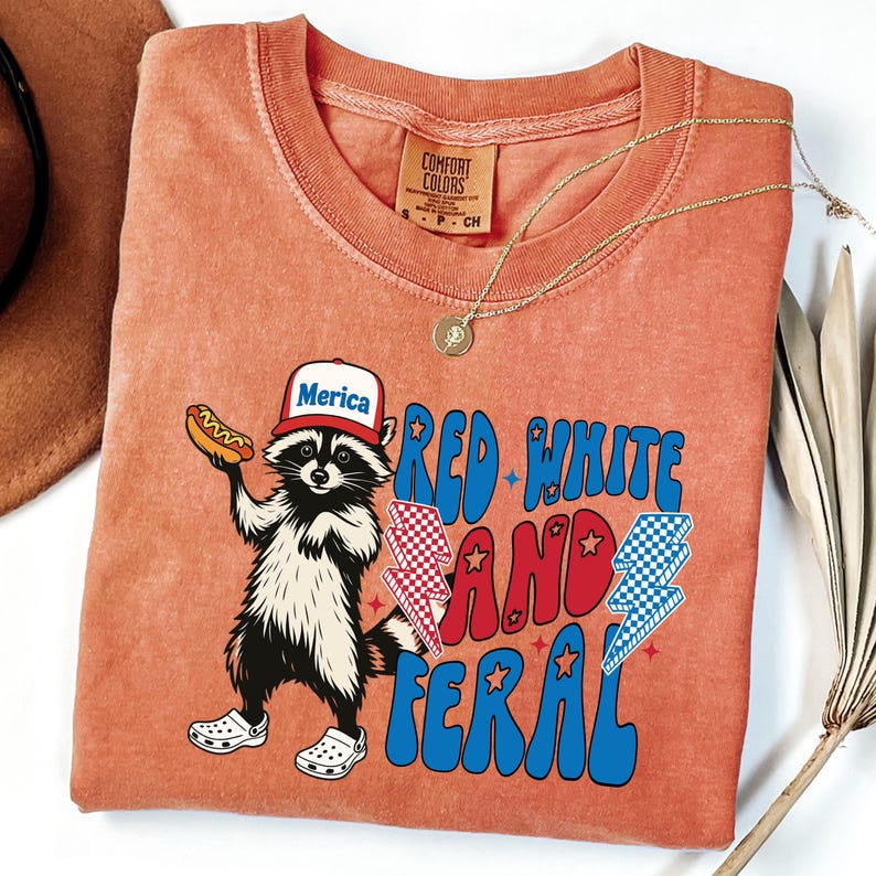 May include: Orange Comfort Colors t-shirt featuring a raccoon wearing a "Merica" hat and holding a hot dog. The shirt has the text "Red White and Feral" in red and blue, with lightning bolt graphics. A gold necklace is draped on the shirt.