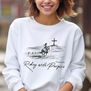 May include: White sweatshirt featuring a black and white illustration of a person riding a horse towards a cross. The text "Riding with Purpose" and "Proverbs 19:21" is printed on the sweatshirt.