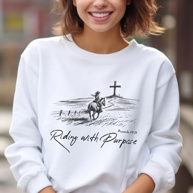 May include: White sweatshirt featuring a black and white illustration of a person riding a horse towards a cross. The text "Riding with Purpose" and "Proverbs 19:21" is printed on the sweatshirt.